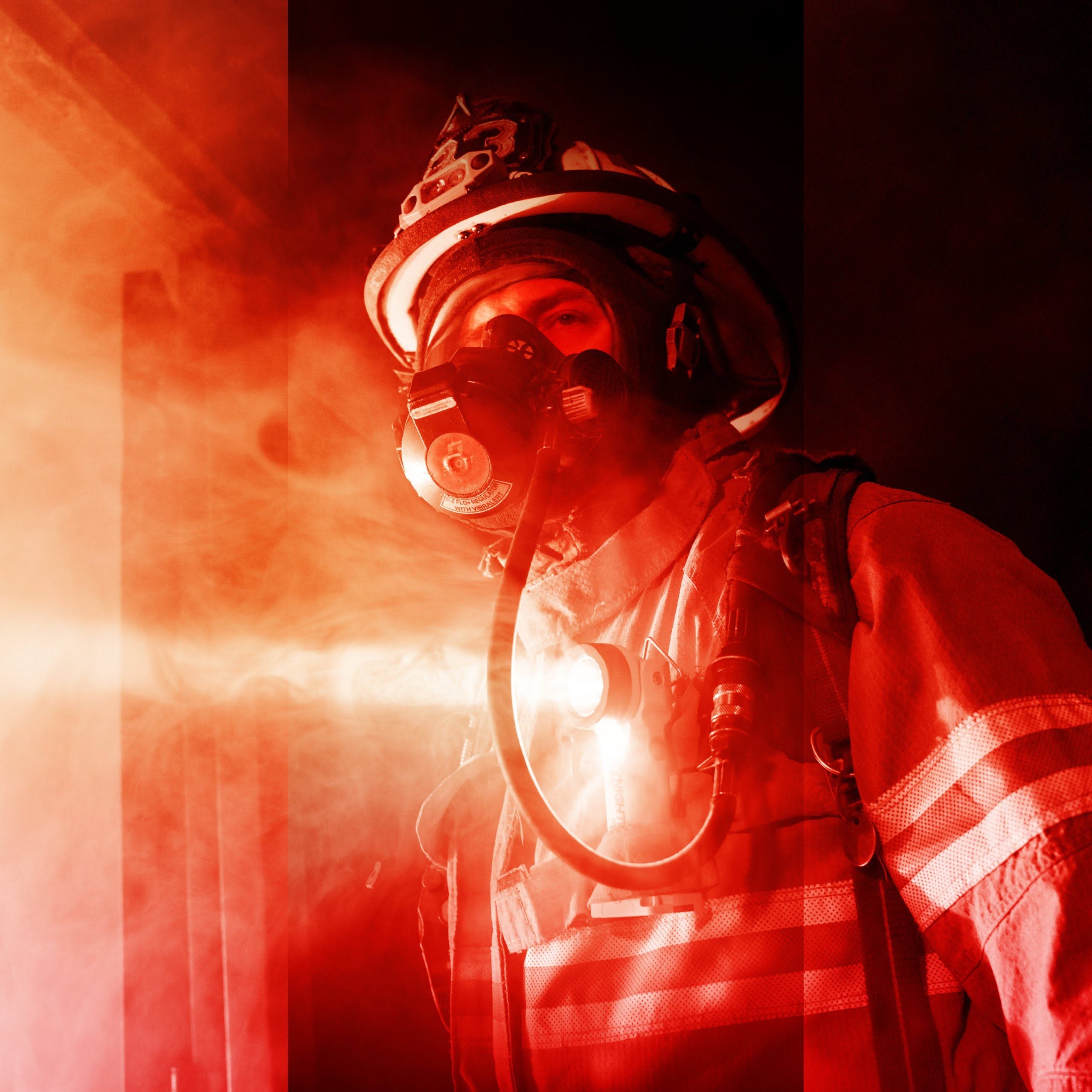 A firefighter in full gear and breathing apparatus stands in a smoky, red-lit environment, using the Nightstick XPP-5566GX INTRANT® Intrinsically Safe Dual-Light™ Angle Light (3 AA) to navigate hazardous conditions.