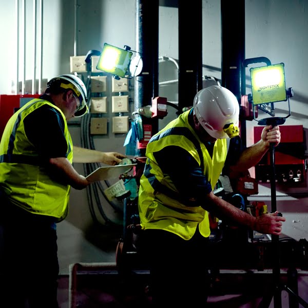 Two workers in safety vests and helmets inspect electrical panels and adjust the Nightstick XPR-5592GCX Intrinsically Safe Rechargeable LED Scene Light Kit, ensuring reliable lighting in hazardous industrial environments.
