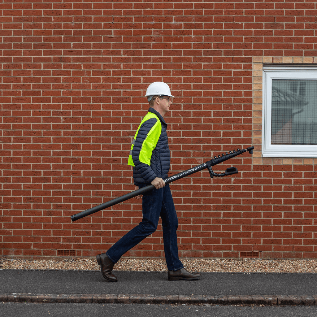 Wearing a hard hat and high-visibility jacket, a person walks by a brick wall, carrying the Armadex Inspection Kit OZC with a 35-foot mast and Android device for rugged, high-reach inspections.