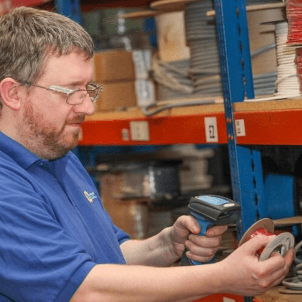 A man wearing glasses scans a spool of wire with an Extronics iSCAN211 Series Zone 1 Handheld Bluetooth Barcode Scanner in a storage area filled with shelves of supplies.