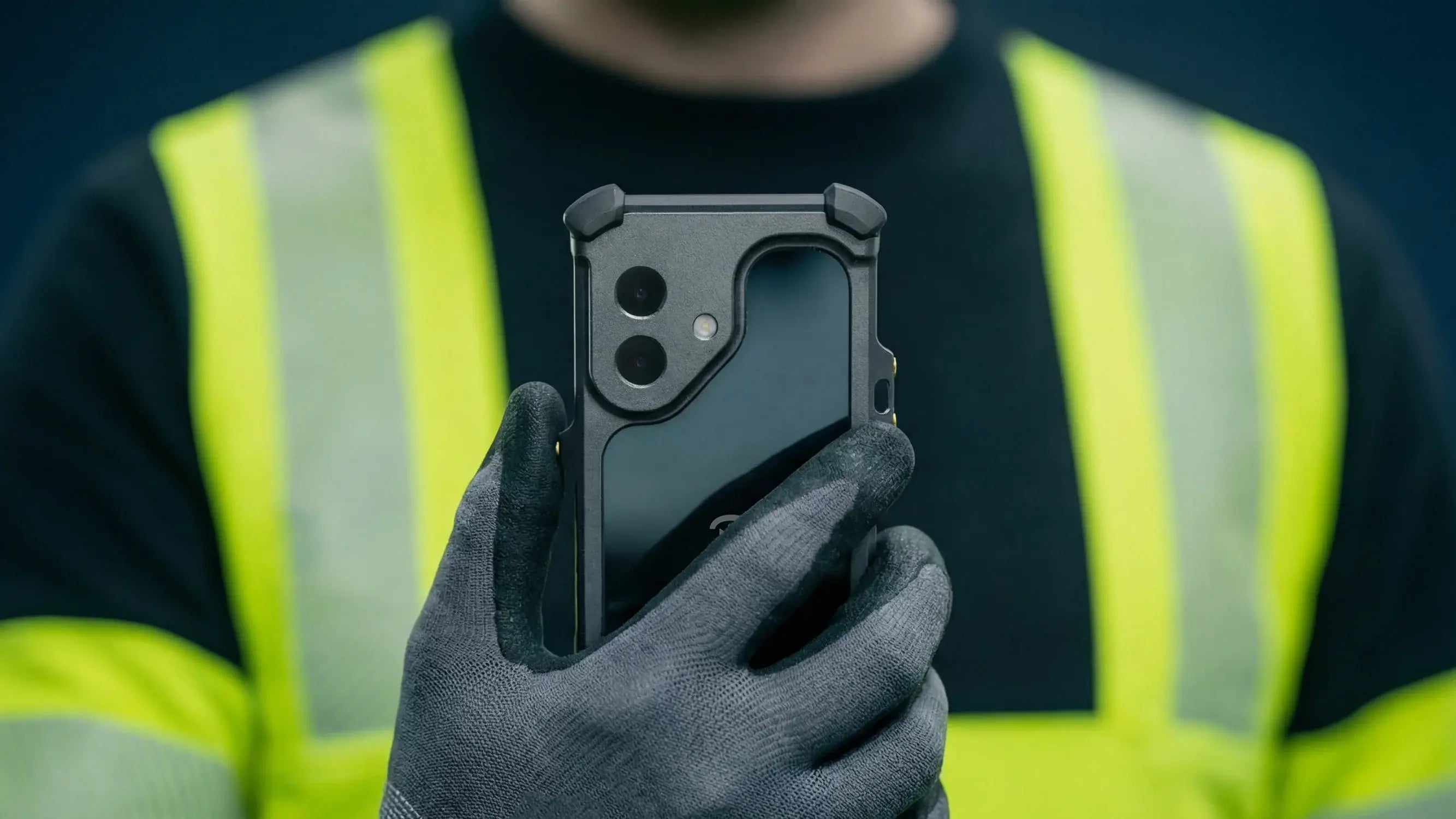 A person in a yellow safety vest and gloves holds up an iPhone 17 with the Xshielder ATEX & IECEx Zone 1/21 Cover, highlighting the case and camera. The phone is included; background is blurred.