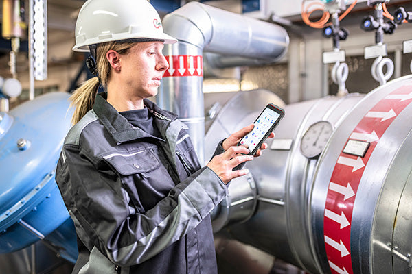 A worker in safety gear uses the i.safe MOBILE IS540.1 Intrinsically Safe (C1/D1) Smartphone with Dual-SIM to access data on a CBRS private cellular network in an industrial facility, large pipes and valves visible in the background.