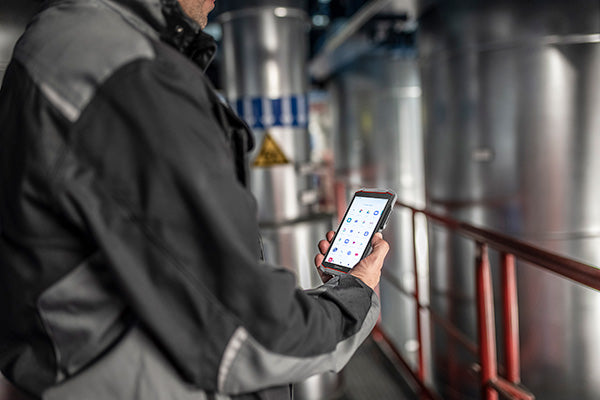 A worker in industrial gear uses the i.safe MOBILE IS540.1 Intrinsically Safe (C1/D1) Smartphone in a facility with pipes and warning signs, showcasing its CBRS private cellular connectivity for safe, seamless operations.