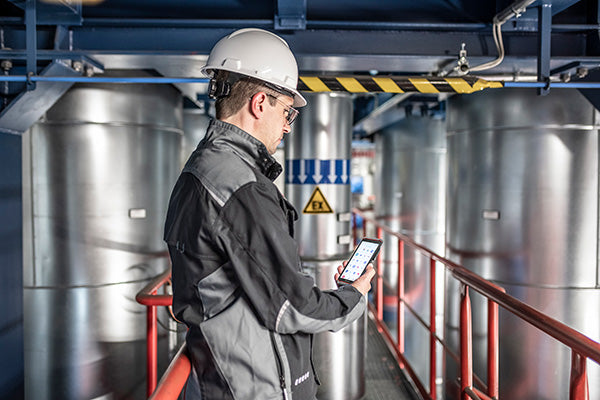 A man in a hard hat and safety glasses uses an i.safe MOBILE IS540.1 Intrinsically Safe (C1/D1) Smartphone with Dual-SIM in an industrial facility surrounded by large metal pipes and warning signs.